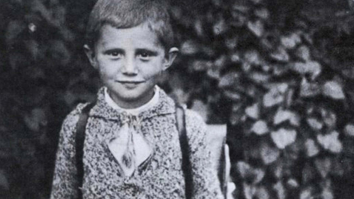 Schwarzweiß-Foto eines Jungen mit Schulranzen, der in die Kamera lächelt.
