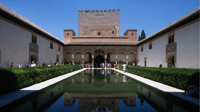 Innenhof in der Alhambra von Granada. Ein Turm und die Bögen des Innenhofes spiegeln sich in einem länglichen Wasserbecken.