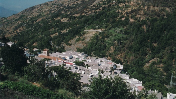 Blick von oben auf ein Dorf, das an einem Hang liegt. Man sieht graue Flachdächer, aus denen kleine, weiße Schornsteine herausschauen und eine Kirche im Zentrum des Dorfes.
