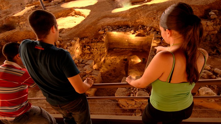 Zwei Besucher stehen an einer Brüstung und blicken in eine Höhle hinab. Unten ist eine Ausgrabungsstätte mit hellbraunen Steinen zu sehen.