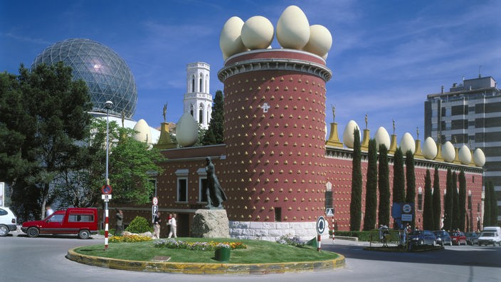 Blick von außen auf das 'Theatre-Museu Dali'. Im Vordergrund ein roter Turm, rechts und links daneben abgehende rote Fassaden des Gebäudes. Auf dem Turm und den Seitenflügeln stehen zahlreiche überdimensionale Eier.