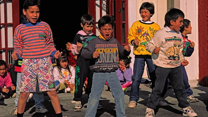 Das Bild zeigt sieben Maori-Kinder, die einen Tanz aufführen. Im Hintergrund sitzen weitere Maori-Kinder auf dem Boden.