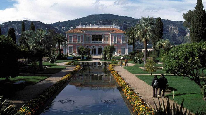 Das Farbfoto zeigt eine zweigeschossige herrschaftliche Villa im Stil von 1900 mit einem geometrisch angelegten Garten. Davor liegt ein künstlicher Wassergraben, umgeben von Blumenbeeten.