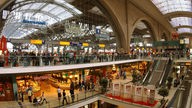 Einkaufszentrum des Leipziger Bahnhofs sind drei Etagen zu erkennen, die durch Rolltreppen miteinander verbunden sind. Auf den einzelnen Etagen sieht man große Menschengruppen.