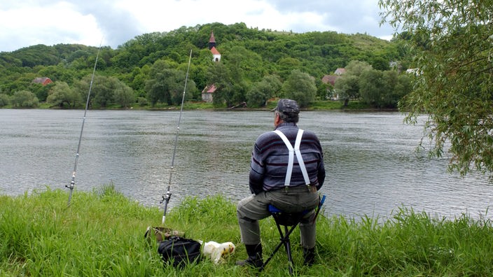 Ein Angler sitzt im Nationalpark Unteres Odertal an der Oder. Neben ihm stehen zwei Angeln und sein Fahrrad. Am gegenüberliegenden Flussufer steht eine Kapelle.