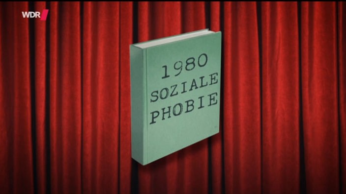 Ein grünes Buch vor einem roten Vohang auf dem steht '1980 Sozial Phobie'.