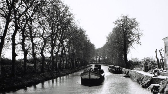 Das Schwarzweiß-Foto zeigt einen gradlinigen Kanal, dessen Ufer Wege mit Bäumen gesäumt sind. Ein Schiff befährt den Kanal. Eines liegt am Ufer.