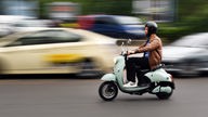 Mann fährt auf grünem Elektro-Roller (Typ Vespa) im Straßenverkehr 