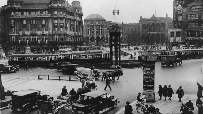 Schwarzweiß-Foto: Der Verkehr am Potsdamer Platz in den 20er Jahren. In einem großen Kreisverkehr sind Autos, Straßenbahnen und Pferdekutschen zu sehen.