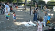 Nachwuchsmusiker spielen auf Instrumenten und singen am Eingang eines Parkes. Einige Passanten schauen zu.