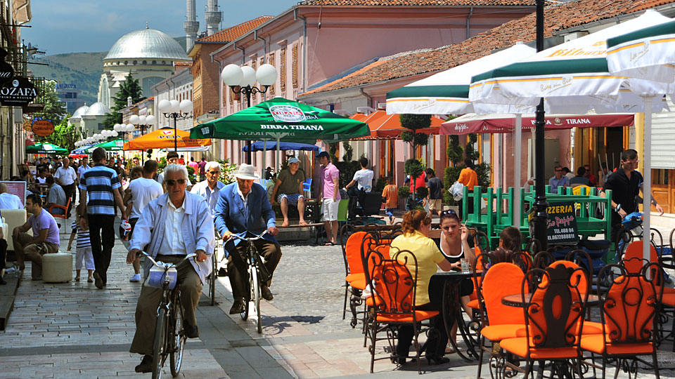 Platz mit Fahrradfahrern in der Fußgängerzone, verschiedene dicht nebeneinander gelegene Cafés mit bunten Stühlen und Sonnenschirmen.
