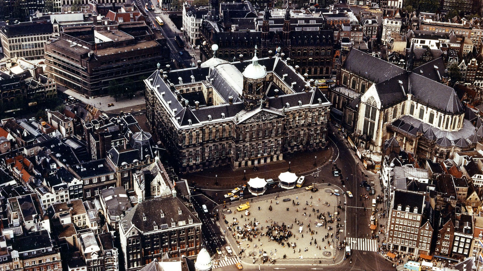 Das Bild, eine Luftaufnahme, zeigt den historischen Marktplatz von Amsterdam mit dem Königlichen Palast. Auf dem Platz sind viele Menschen zu erkennen