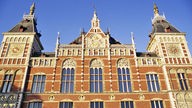 Das Bild zeigt die verzierte Fassade des Amsterdamer Hauptbahnhofs aus der Froschperspektive