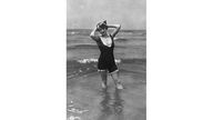 Eine junge Frau mit Hut und schwarzem, ärmellosem Badeanzug mit kurzem Bein und weitem Ausschnitt steht kess im Wasser.