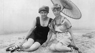 Zwei edel gekleidete Damen sitzen fein am Strand, mit Hüten und einem vornehmen Schirm.