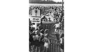 Schwarzweiß-Foto eines vollen Strandbads. In der Mitte steht ein Bademeister mit Sprechrohr und einem Kind auf dem Arm.