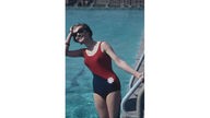 Eine Frau mit Sonnenbrille im blauroten Badeanzug hält sich an einer Leiter am Schwimmbad-Beckenrand fest und schaut in die Sonne.