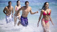 Junge Leute in Badekleidung rennen durch seichtes Wasser.