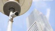 Ein Mast mite einer Überwachungskamera im öffentlichen Raum; im Hintergrund ist ein Hochhaus zu sehen