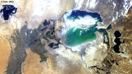 Satellitenaufnahme: In einer vorwiegend braunen Landschaft befindet sich ein See in blauen und grünlichen Farben