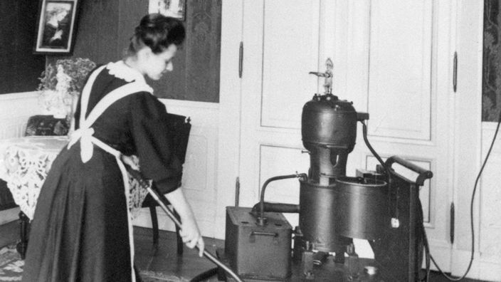Schwarzweißfoto von 1906: Ein Hausmädchen arbeitet mit einer "Entstäubungspumpe", einer Art Staubsauger
