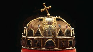 Bild der ungarischen Stephanskrone: Die helmähnliche Krone aus vergoldetem Silber trägt ein schiefes Kreuz und ist mit Dreiecken und Bögen besetzt. Dazwischen befinden sich einige Perlenschnüre.