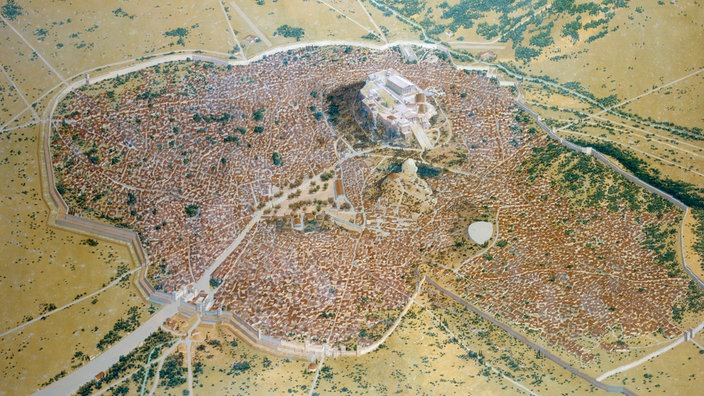 In einer Landschaft mit geringer Vegetation sieht man aus der Vogelperspektive eine Stadt liegen. Auffallend ist ein Hügel innerhalb der Stadt mit einer Tempelanlage. | Bildquelle: AKG/Peter Connolly In einer Landschaft mit geringer Vegetation sieht man aus der Vogelperspektive eine Stadt liegen. Auffallend ist ein Hügel innerhalb der Stadt mit einer Tempelanlage.