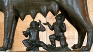 Unter einer stilisierten Wölfin aus Bronze sitzen zwei Säuglinge und trinken an den Zitzen des Tieres.