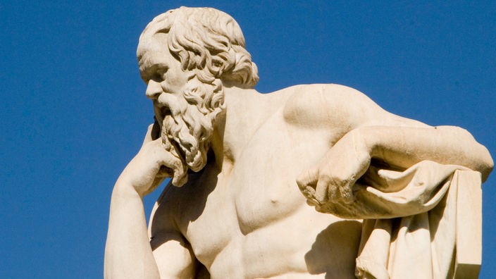 Eine Sitzstatue aus hellem Marmor in Athen. Sokrates sitzt nachdenklich nach vorne gebeugt. Im Hintergrund der Statue ist der blaue Himmel zu sehen. 