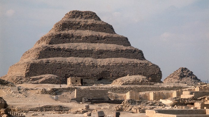 Blick auf eine Pyramide in Stufenform. Vor der Pyramide sind Grabungsfelder zu sehen.