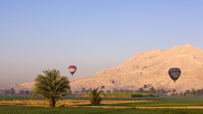 Blick über den Nil auf ein paar Häuser am gegenüber liegenden Ufer. Im Hintergrund hohe, stufenförmige Berge. Am blauen Himmel ist ein roter Heißluftballon.