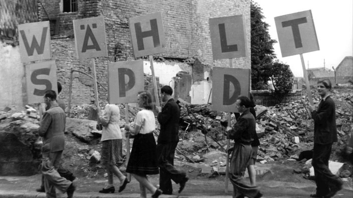 Vor den Trümmern eines Hauses tragen junge Männer und Frauen Schilder, die den Schriftzug "Wählt SPD" ergeben, Juni 1946 | Bildquelle: WDR / dpa Vor den Trümmern eines Hauses tragen junge Männer und Frauen Schilder, die den Schriftzug "Wählt SPD" ergeben, Juni 1946