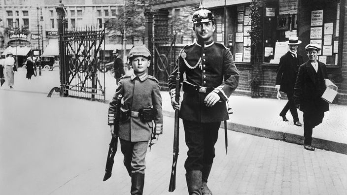 Ein stolzer Sohn in Kinder-Uniform begleitet seinen Vater, der in den Krieg zieht, zum Bahnhof