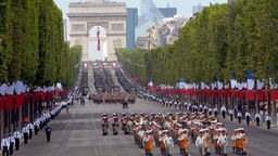 Vor dem Arc de Triomphe in Paris marschieren Legionäre der Fremdenlegion auf die Kamera zu. Die breite Straße ist gesäumt von grünen Bäumen und französischen Flaggen.