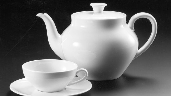Vor schwarzem Hintergrund stehen eine schlichte weiße Teekanne und eine Tasse. Beide sind sehr unauffällig gestaltet.