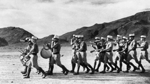 Ein Trupp uniformierter Fremdenlegionäre marschiert musizierend durch karges Gelände.