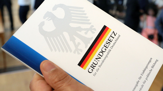 Eine Hand hält das Grundgesetz in die Kamera, auf dem Titelblatt der Bundesadler, im Hintergrund eine Deutschlandfahne.
