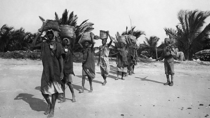 Deutsch-Ostafrika: Gefangene in Ketten tragen Lasten an einem Aufseher vorbei | Bildquelle: picture alliance / ullstein bild (Haeckel Archiv) Deutsch-Ostafrika: Gefangene in Ketten tragen Lasten an einem Aufseher vorbei