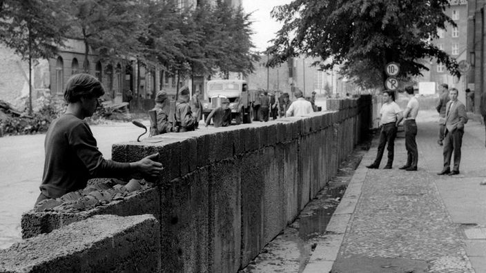 1961: Im Vordergrund schichtet jemand Steine auf die neu entstehende Mauer, weiter hinten schauen Bürger und Soldaten zu