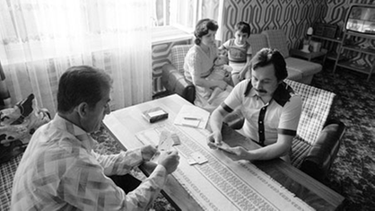 Türkische Gastarbeiter-Familie in den 70ern. Zwei Männer sitzen im Wohnzimmer an einem Tisch und spielen Karten. Eine Frau mit Kind sitzt im Hintergrund auf dem Sofa.