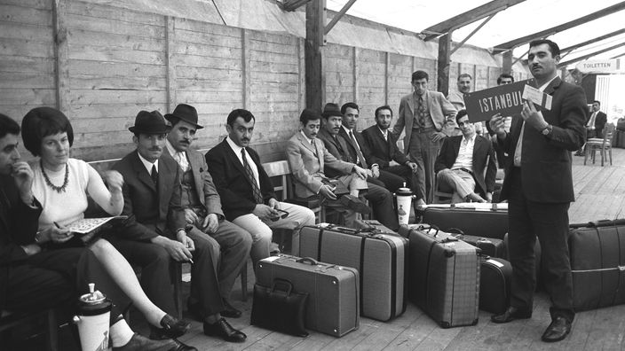 Mehrere Männer und eine Frau sitzen mit gepackten Koffern in einer Wartehalle. Einer der Männer hält ein Schild mit der Aufschrift 'Istanbul'.