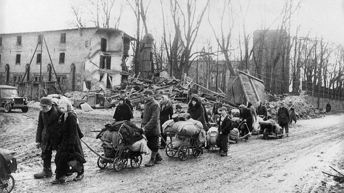 Archivbild: Mehrere Männer, Frauen und Kinder ziehen mit Handkarren durch ein Dorf, im Hintergrund zerstörte Gebäude