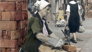 Koloriertes Foto einer Frau mit Kopftuch. Sie sitzt auf einem Stapel Ziegelsteine und bearbeitet mit einem Werkzeug einen Stein.