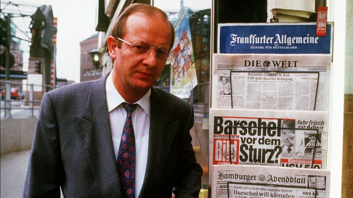 Medienreferent Reiner Pfeiffer posiert neben Zeitungsständer mit Barschel-Aufmachern.