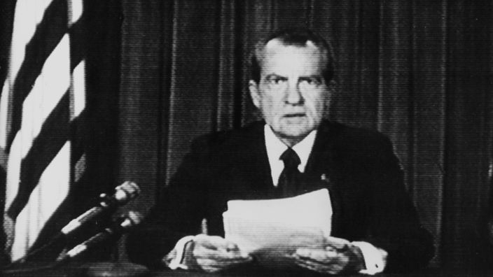 Richard Nixon sitzt an einem Tisch, ein Blatt Papier in seinen Händen, neben sich die amerikanische Flagge und blickt ernst in die Kamera.