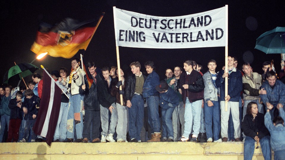 Berliner stehen mit Flaggen und Plakten auf der Berliner Mauer am Brandenburger Tor.