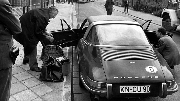 Polizisten schauen zu, wie zwei Männer einen dunklen Porsche 911 durchsuchen. Die Türen sind offen, auf dem Bürgersteig stehen mehrere Taschen. | Bildquelle: dpa Polizisten schauen zu, wie zwei Männer einen dunklen Porsche 911 durchsuchen. Die Türen sind offen, auf dem Bürgersteig stehen mehrere Taschen.