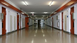 Ein langer Gefängnisgang mit blank gewienertem Boden. Nach links und rechts gehen zahlreiche Zellen ab, die mit dicken roten Stahltüren verschlossen sind.