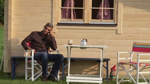 Ein Mann sitzt vor einem Holzhäuschen auf einem Stuhl.