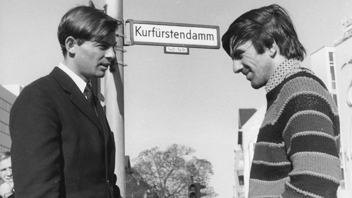 Rudi Dutschke diskutiert mit einem Freund. Er trägt einen gestreiften Pullover.